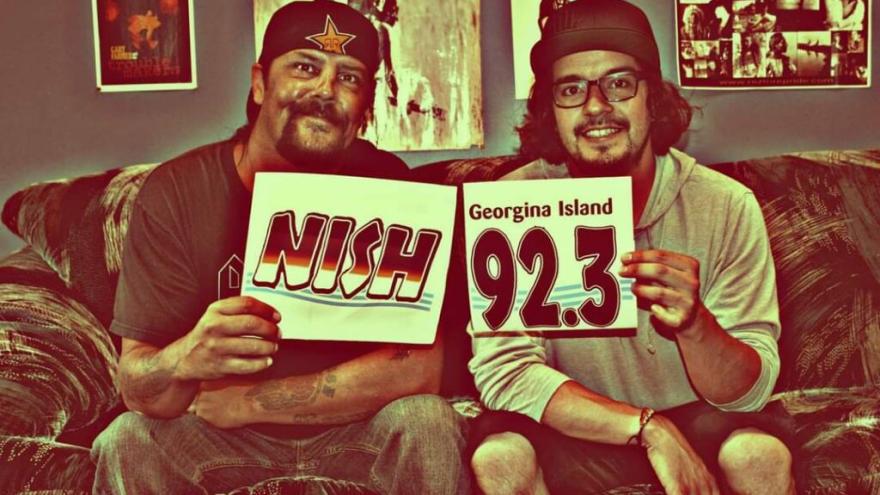 Two smiling men holding radio station signs while sitting on a sofa.
