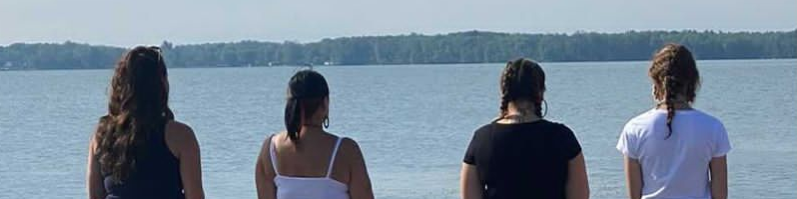 Four young women stand by a lake, facing the water.