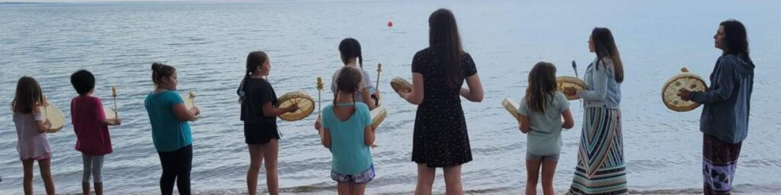 Young people stand on a beach holding drums, facing the sea.