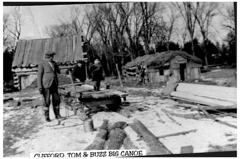 Man stands outside rustic cabins in a snowy rural setting.