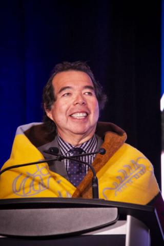 Man smiling at podium, wearing yellow blanket with blue and brown patterns.