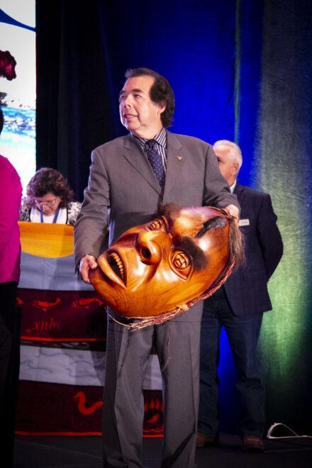 Bill McCue holding a large carved wooden mask on stage.