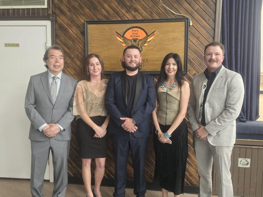 Five people standing in a row and smiling, dressed in formal attire.