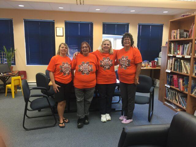 Four women in matching orange shirts stand together in a library.