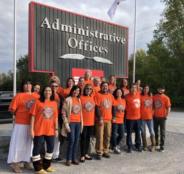 Group wearing orange shirts standing outside by a sign for Administrative Offices.